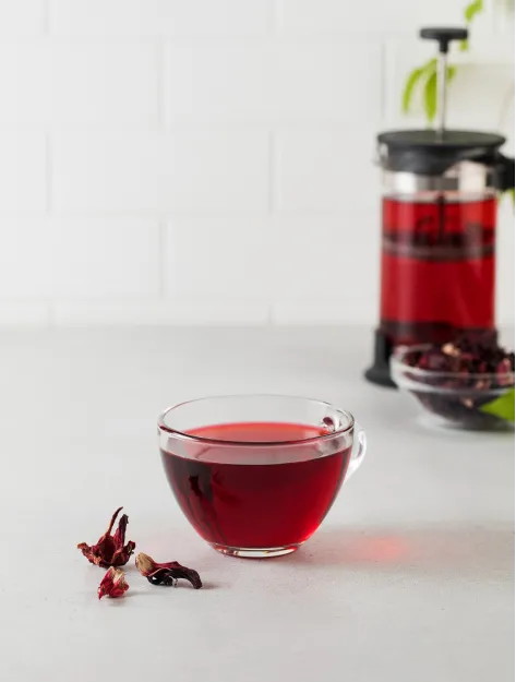 A glass cup of red hibiscus tea next to a French press and dried hibiscus flowers.